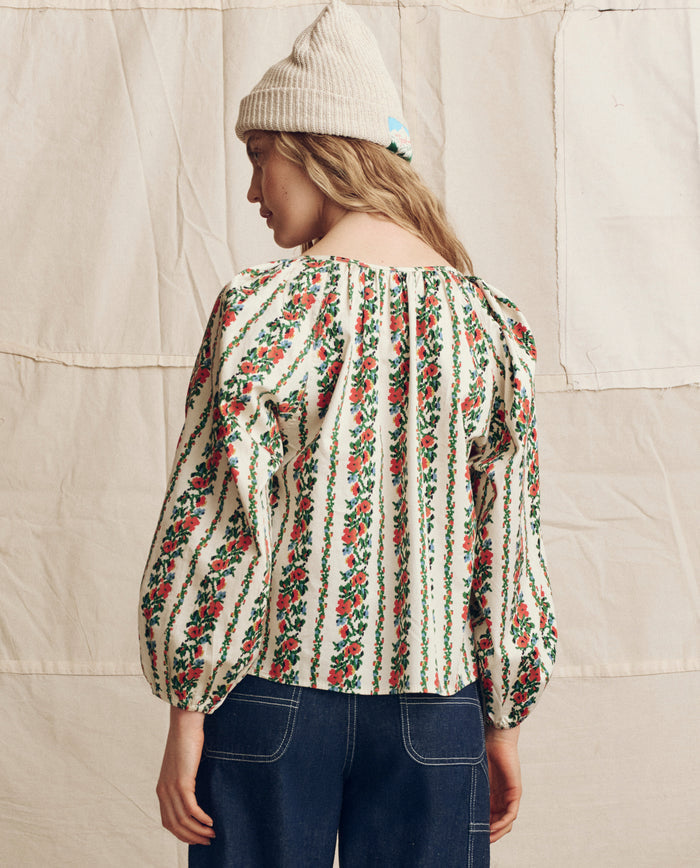 A person stands facing away against a light fabric backdrop, wearing THE GREAT. PS26’s The Chariot Top in Tapestry Floral—a puffed-sleeve blouse—paired with a cream knit beanie and dark blue jeans.