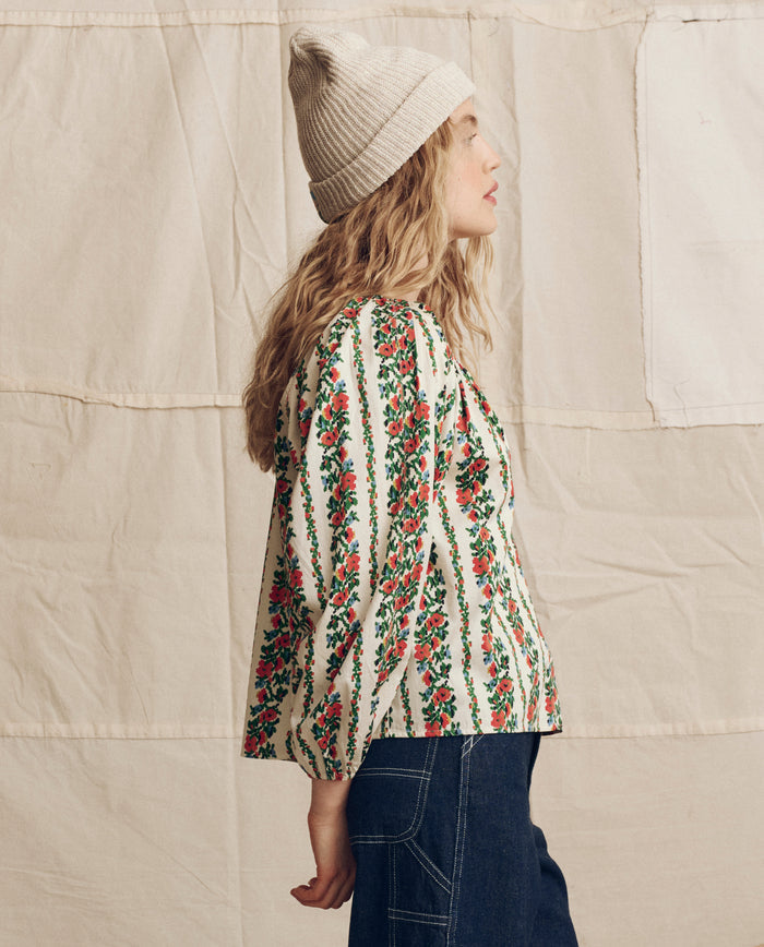 A person in THE GREAT. PS26 The Chariot Top -- Tapestry Floral stands in profile, wearing a cream knit beanie and dark jeans against a beige fabric backdrop.