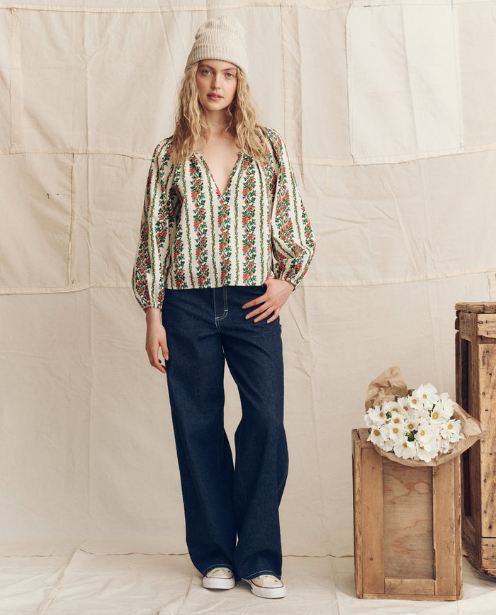 A woman poses indoors beside wooden crates of white flowers, wearing THE GREAT. PS26 The Chariot Top in Tapestry Floral, paired with wide-leg jeans, white sneakers, and a knit hat against a neutral fabric backdrop.