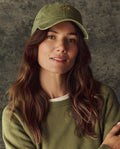 A woman with long brown hair wears a green sweater and The Classic Baseball Hat -- Army by THE GREAT. H25 BRIGHT FLEECEBACK, featuring vintage-style embroidery and an adjustable back, standing before a textured gray background.