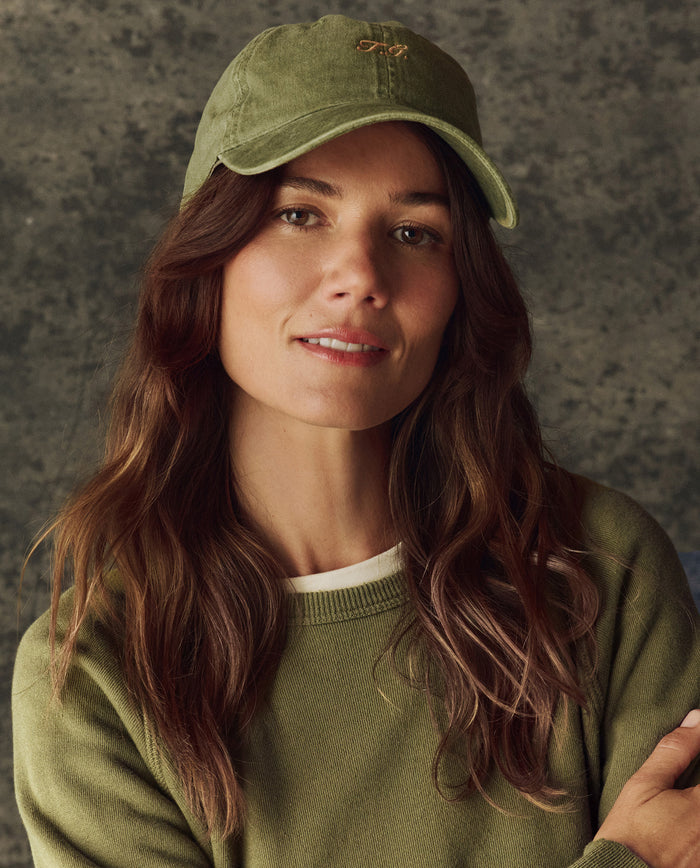 A woman with long brown hair wears a green sweater and The Classic Baseball Hat -- Army by THE GREAT. H25 BRIGHT FLEECEBACK, featuring vintage-style embroidery and an adjustable back, standing before a textured gray background.