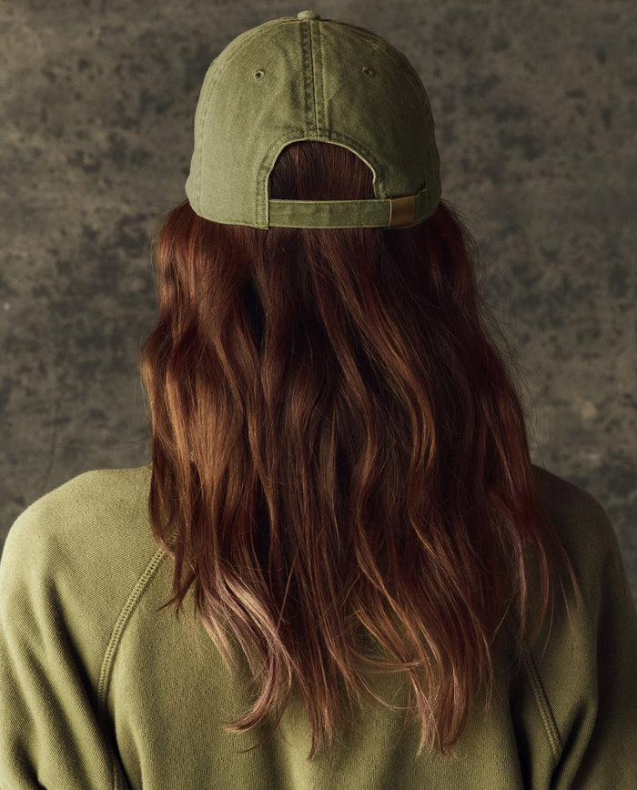 A person with long brown hair is seen from behind wearing THE GREAT. H25 BRIGHT FLEECEBACK Classic Baseball Hat in Army, paired with a matching sweatshirt, against a textured gray backdrop.