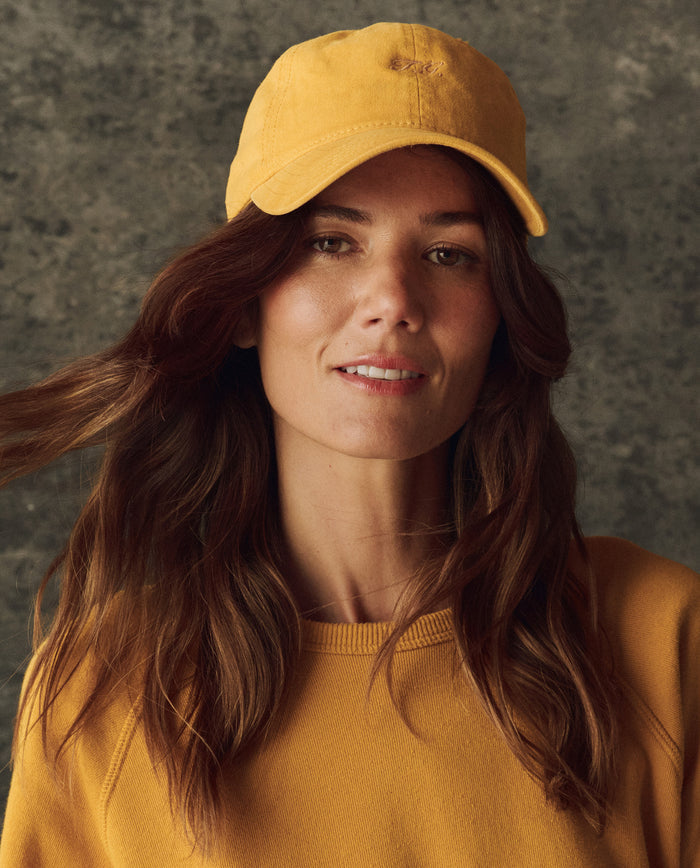 A woman with long brown hair wears THE GREAT. H25 BRIGHT FLEECEBACK yellow sweatshirt and The Classic Baseball Hat – Bright Yellow, posing against a textured gray background.