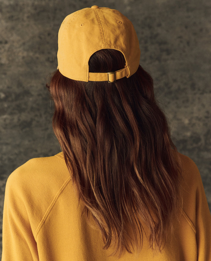 A person with long brown hair, seen from behind against a textured gray background, wears THE GREAT. H25 BRIGHT FLEECEBACK's The Classic Baseball Hat in bright yellow with a matching top.