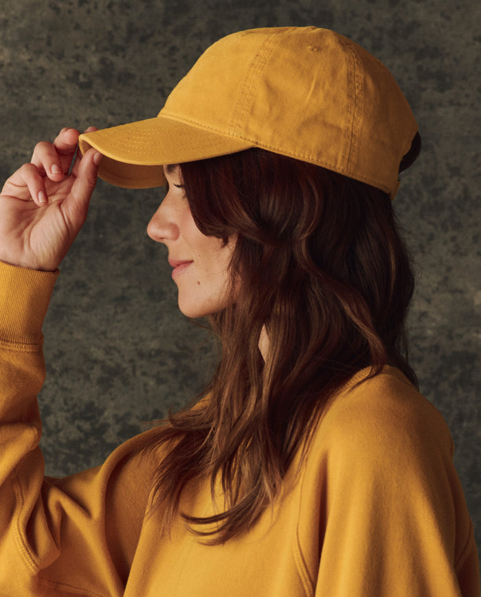 A person with long brown hair wears THE GREAT. H25 BRIGHT FLEECEBACK's The Classic Baseball Hat in bright yellow and a matching sweatshirt, shown in profile against a textured dark background.
