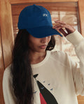 A person wearing The Baseball Hat in cobalt by THE GREAT. SU26 and a cream sweatshirt with an abstract bird design.