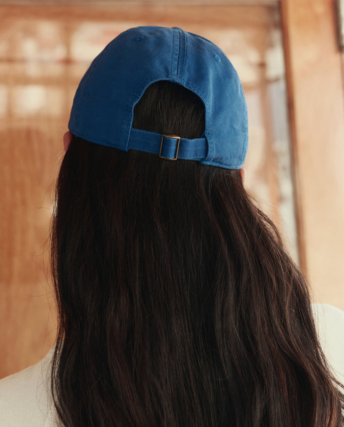 Person with long, dark hair wears THE GREAT. SU26's The Baseball Hat in Cobalt backward indoors, facing away from the camera.