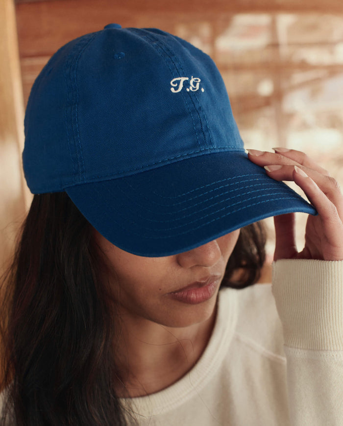 A person wears The Baseball Hat in Cobalt by THE GREAT. SU26, featuring embroidered "T.G." initials on the front, partially covering their face and paired with a light-colored top for a casual touch.