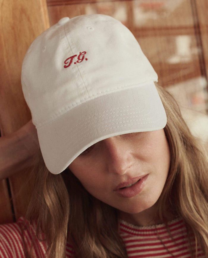 A person wearing The Baseball Hat in white by THE GREAT. SU26, featuring red "T.G." embroidery, paired with a red and white striped top.