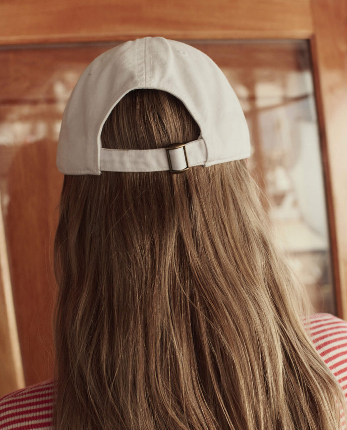A person with long brown hair wears THE GREAT. SU26’s The Baseball Hat in white backwards while standing indoors near a wooden surface.