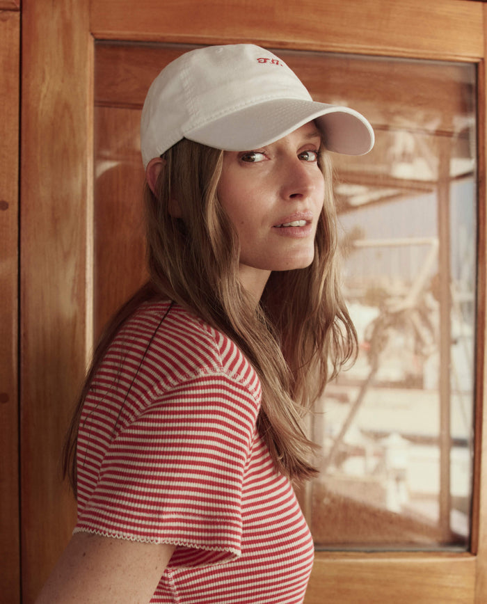 A woman in a red-and-white striped shirt and THE GREAT. SU26 The Baseball Hat in White stands indoors by a wooden-framed glass door, facing the camera.