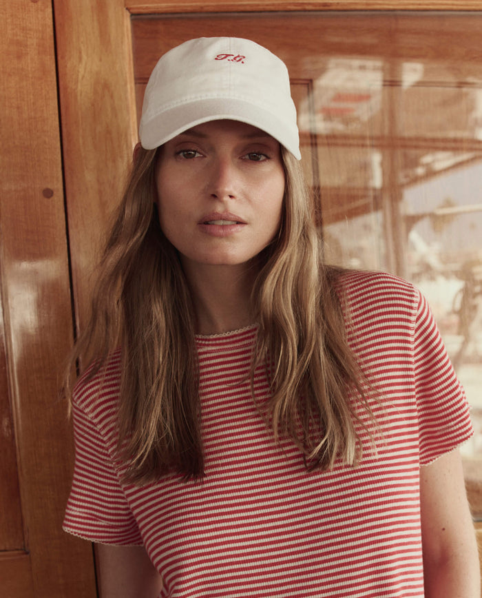 A woman with long light brown hair wears THE GREAT. SU26 The Baseball Hat in white and a red-and-white striped T-shirt, standing in front of a wooden wall.