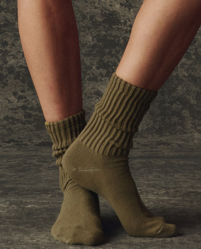 A person stands on tiptoes wearing THE GREAT. H25 BRIGHT FLEECEBACK’s The Classic Crew Sock – Army, shown in olive green against a textured dark background.