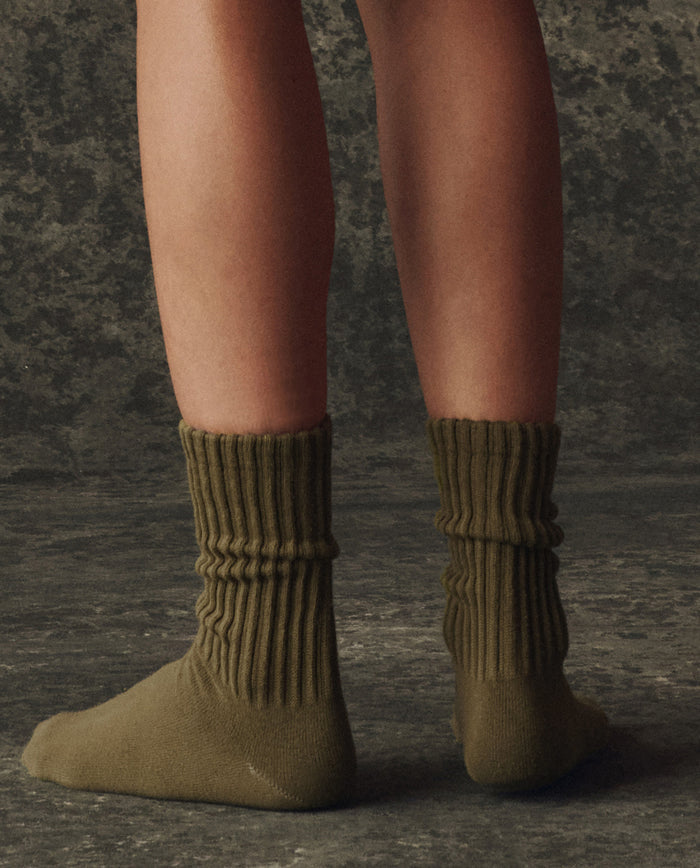 A person stands on a dark textured floor, shown from the knees down, wearing THE GREAT. H25 BRIGHT FLEECEBACK's The Classic Crew Sock -- Army, crafted from soft cotton yarn with a ribbed design.