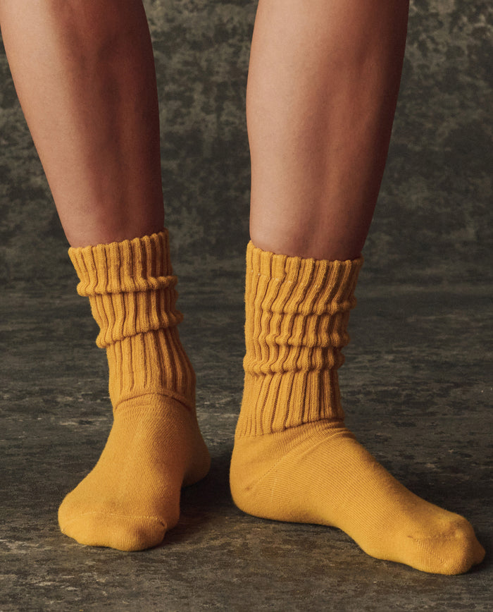 A person wears THE GREAT. H25 BRIGHT FLEECEBACK The Classic Crew Sock in Bright Yellow, standing on a textured dark surface, with only their lower legs and feet visible.
