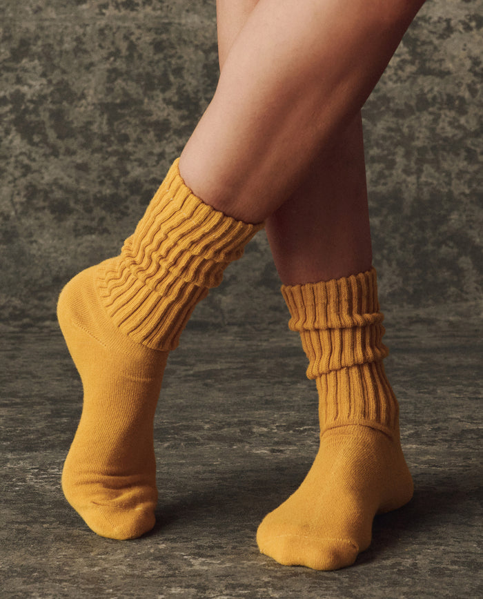 A person in The Classic Crew Sock – Bright Yellow by THE GREAT. H25 BRIGHT FLEECEBACK stands on a textured dark surface, feet slightly crossed in a vintage athletic style.