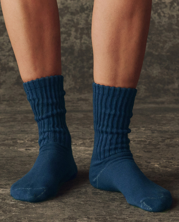 A person wearing THE GREAT. H25 BRIGHT FLEECEBACK The Classic Crew Sock in Navy stands on a dark textured surface.