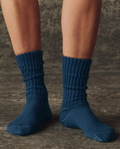 A person wearing THE GREAT. H25 BRIGHT FLEECEBACK The Classic Crew Sock in Navy stands on a dark textured surface.