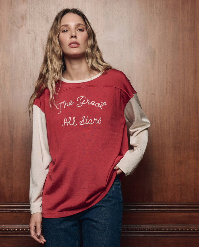A woman with long wavy hair wears THE GREAT. F26 COLLECTORS The Collectors Sport Tee in True Red with Cream, which features "The Great All Stars" lettering, paired with blue jeans as she stands against a wood-paneled wall.