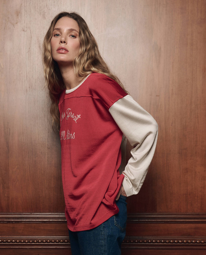 A person with long wavy hair wears THE GREAT. F26 COLLECTORS The Collectors Sport Tee in True Red with Cream and jeans, standing in front of a wooden wall.