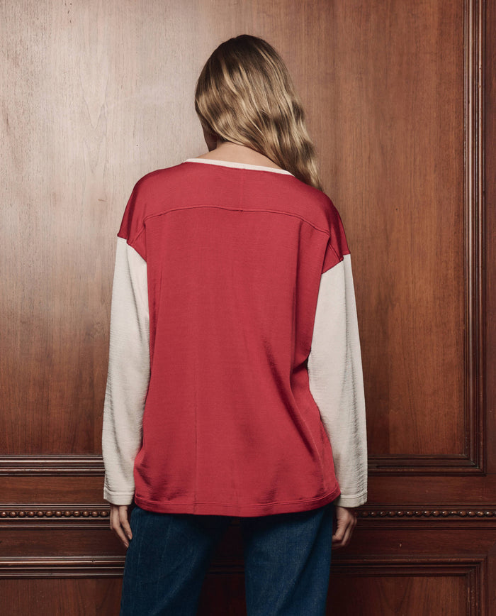 A person with long hair, seen from behind, wearing THE GREAT. F26 COLLECTORS "The Collectors Sport Tee" in True Red with Cream and blue jeans, stands in front of a wooden paneled wall.