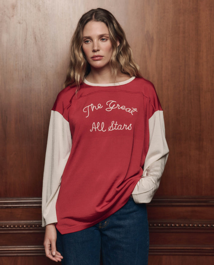 A woman with wavy hair wears THE GREAT. F26 COLLECTORS The Collectors Sport Tee in True Red with Cream, featuring "The Great All Stars" text, paired with blue jeans and standing against a wooden panel background.