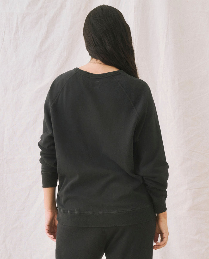 A person with long hair stands facing away, clad in The College Sweatshirt in Solid -- Almost Black by THE GREAT. CORE KNITS and black pants, against a plain, light-colored backdrop.