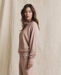 A woman stands sideways against a beige fabric backdrop, looking to the left, wearing THE GREAT. F26 SEASONAL CORE The College Sweatshirt in solid Ballet Pink with matching pants.