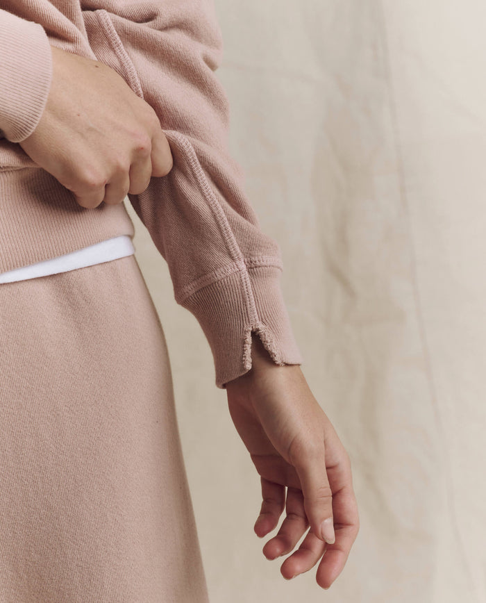 A person wearing THE GREAT.’s The College Sweatshirt. Solid in Ballet Pink from the F26 SEASONAL CORE collection, paired with matching pants, adjusts a sleeve featuring a subtle notch near the cuff. The background remains neutral and softly out of focus, evoking a vintage-inspired style.