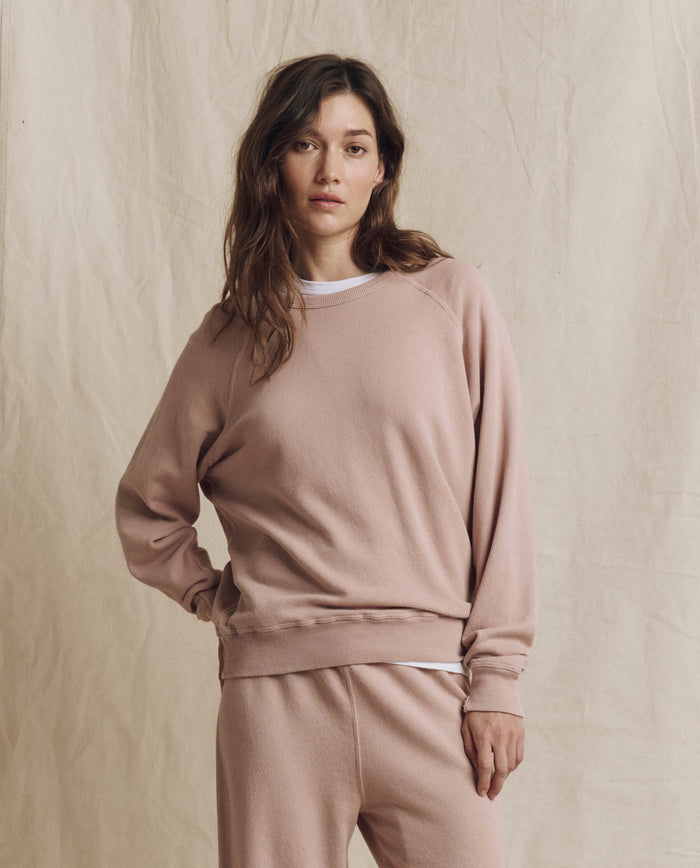 A person wearing THE GREAT. F26 SEASONAL CORE’s The College Sweatshirt in Solid Ballet Pink stands in front of a beige fabric backdrop, looking at the camera.