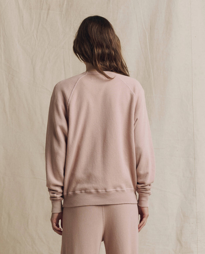 A person stands with their back to the camera, wearing THE GREAT. F26 SEASONAL CORE’s The College Sweatshirt. Solid in Ballet Pink, paired with matching pants against a beige fabric backdrop, embodying effortless style with a touch of handcrafted Los Angeles charm.