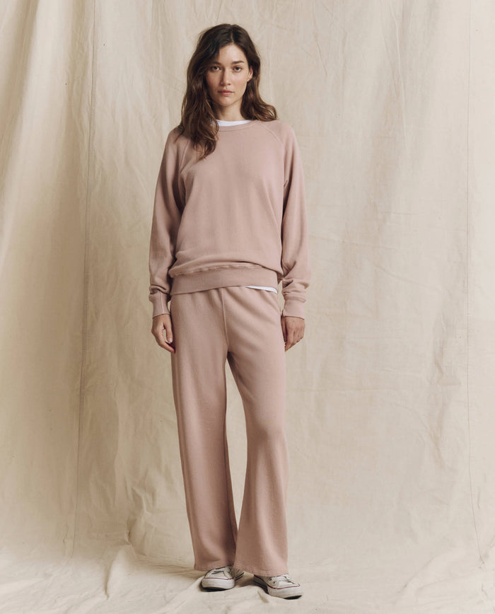 A woman stands in front of a beige fabric backdrop, wearing THE GREAT.’s The College Sweatshirt in Solid Ballet Pink from the F26 SEASONAL CORE collection with matching pants, layered over a white shirt and paired with white sneakers.