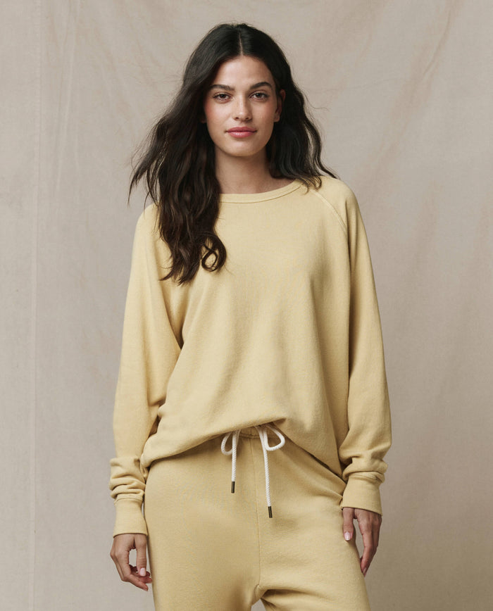 A woman stands facing forward wearing THE GREAT. SP26 SEASONAL CORE The College Sweatshirt in butter yellow against a neutral background.