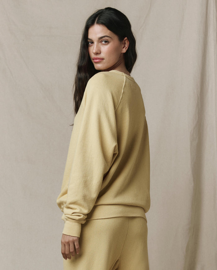 A person with long dark hair wears THE GREAT.'s The College Sweatshirt in Butter Yellow (SP26 SEASONAL CORE), paired with matching pants, standing against a neutral fabric background and looking over their shoulder.