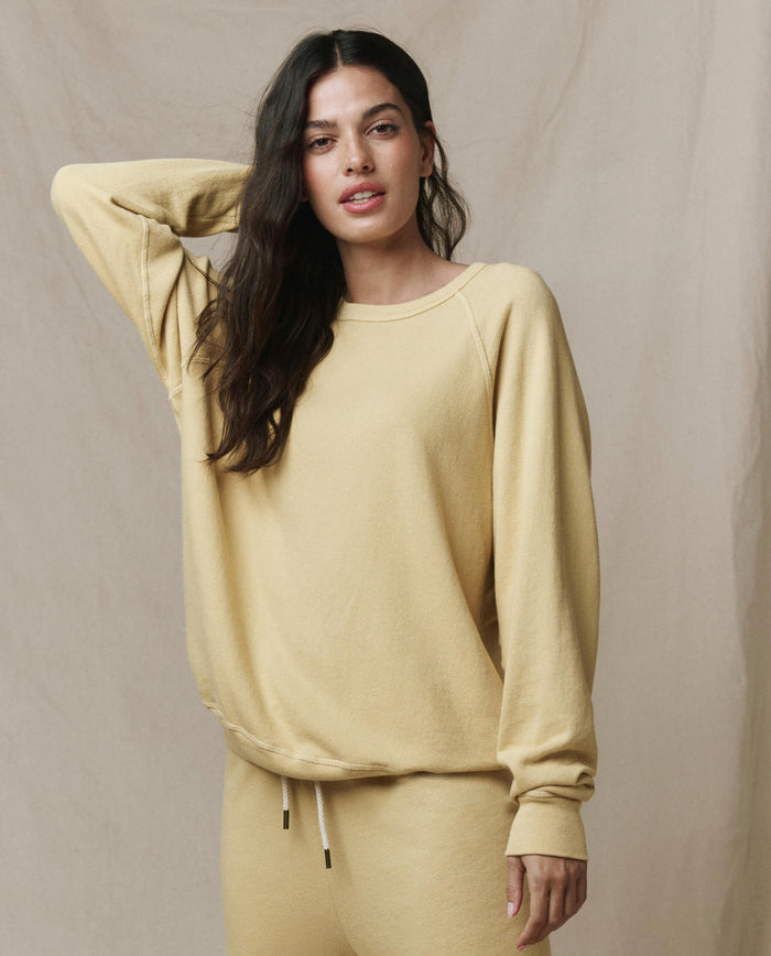 A woman wearing THE GREAT.'s The College Sweatshirt in Butter Yellow (SP26 Seasonal Core) stands against a neutral background, one hand resting on her head.
