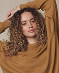 A curly-haired person models THE GREAT.'s The College Sweatshirt in Solid -- Cork (SU26 SEASONAL CORE), arms raised and crossed above their head, looking at the camera against a neutral background.