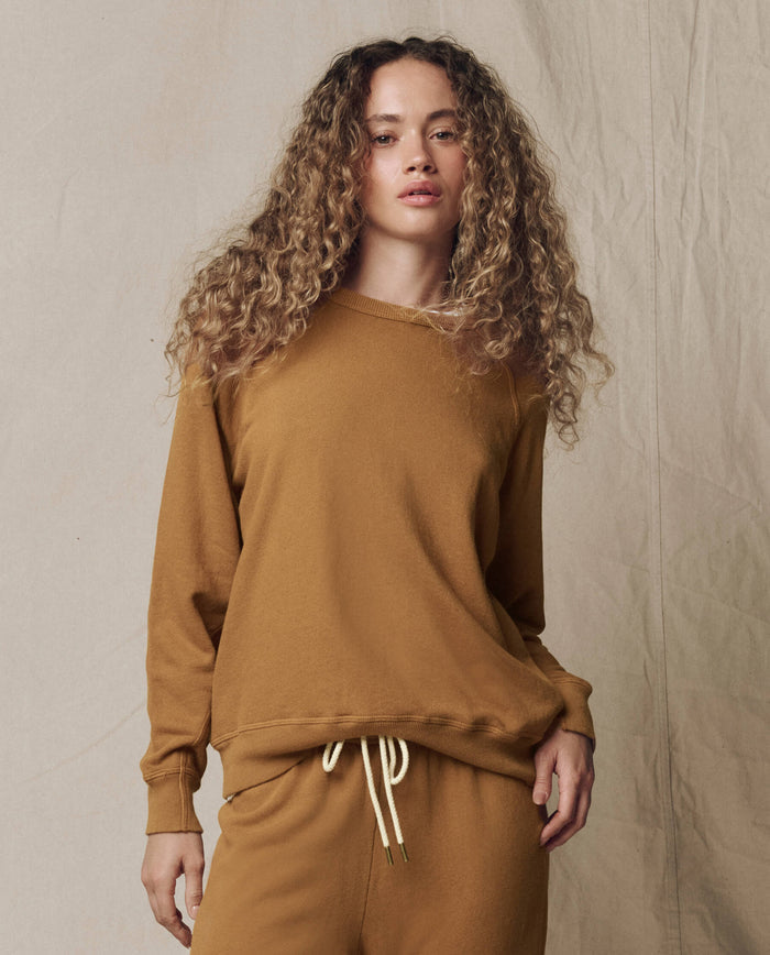 A person with curly hair is wearing THE GREAT. SU26 SEASONAL CORE The College Sweatshirt in Solid -- Cork, standing against a neutral fabric backdrop.