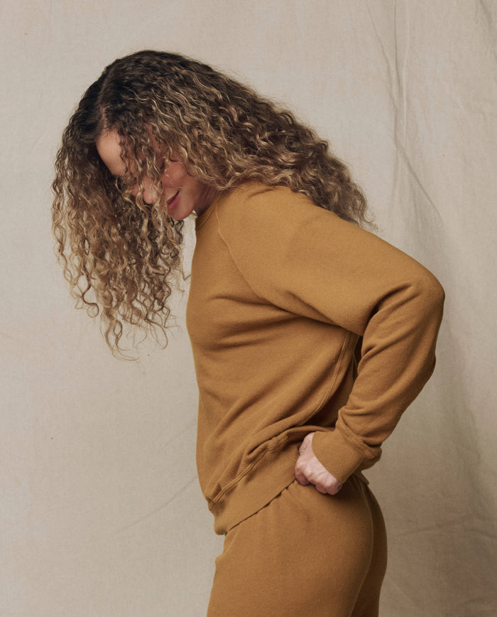 A person with curly hair stands in profile, looking down, wearing THE GREAT. SU26 SEASONAL CORE The College Sweatshirt. Solid -- Cork and matching pants against a plain beige background.