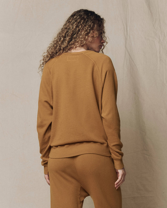 A person with curly hair wears THE GREAT.'s The College Sweatshirt, Solid -- Cork (SU26 SEASONAL CORE), paired with matching sweatpants, standing against a neutral fabric background.