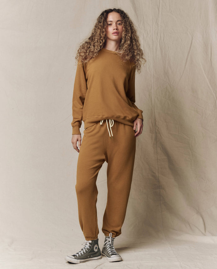 Someone with curly hair wears THE GREAT. SU26 SEASONAL CORE The College Sweatshirt in Solid -- Cork with matching sweatpants and gray high-top sneakers, standing against a beige fabric backdrop.