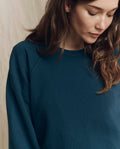 A woman with long brown hair wears THE GREAT. F26 SEASONAL CORE The College Sweatshirt in Solid -- Dark Slate, shown from the shoulders up against a neutral background, looking downward.