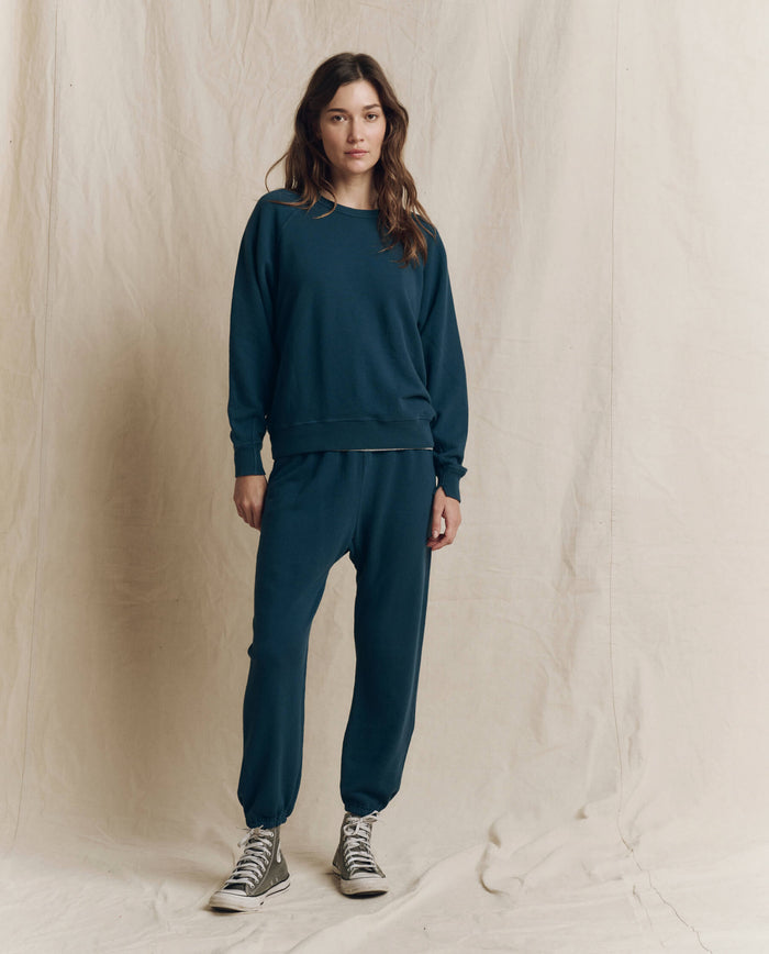 A person stands in front of a beige backdrop, wearing THE GREAT.’s The College Sweatshirt in Solid -- Dark Slate from the F26 SEASONAL CORE collection, paired with matching sweatpants and grey high-top sneakers, effortlessly embodying handcrafted Los Angeles style.