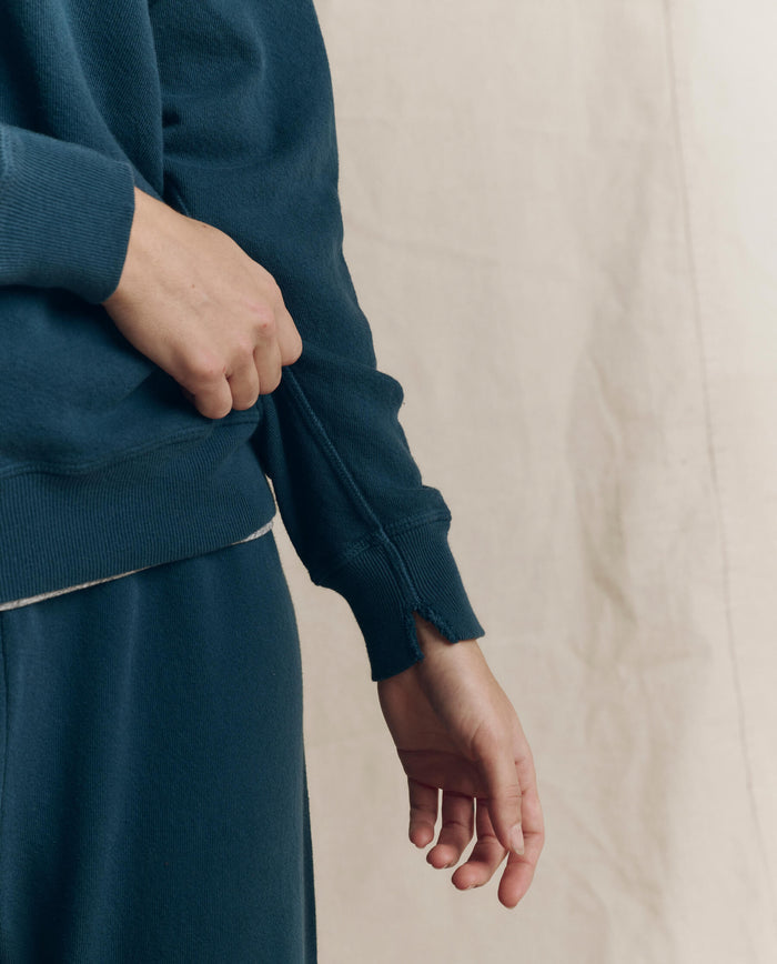 A person wearing THE GREAT. F26 SEASONAL CORE The College Sweatshirt in Solid -- Dark Slate is shown from the shoulders down, with one hand pulling on the sleeve of the vintage-inspired sweatshirt.