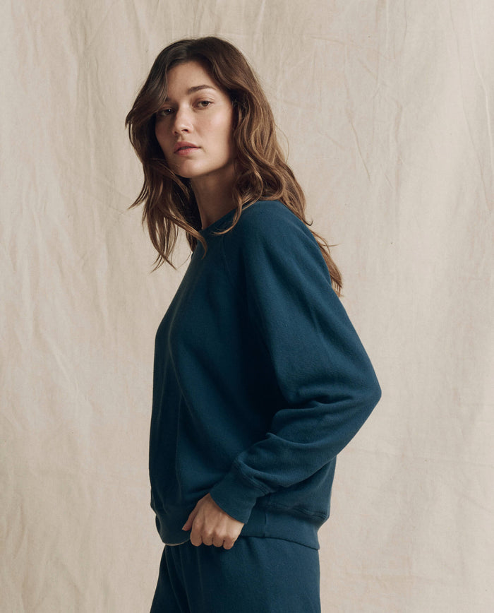 Wearing THE GREAT. F26 SEASONAL CORE’s The College Sweatshirt in Solid -- Dark Slate, a person with long brown hair stands in front of a beige fabric backdrop, embodying the effortless style of handcrafted Los Angeles fashion.
