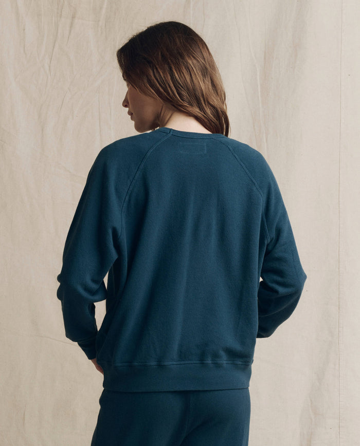A person with brown hair stands facing away from the camera against a beige fabric backdrop, wearing THE GREAT. F26 SEASONAL CORE’s The College Sweatshirt in Solid Dark Slate paired with pants.