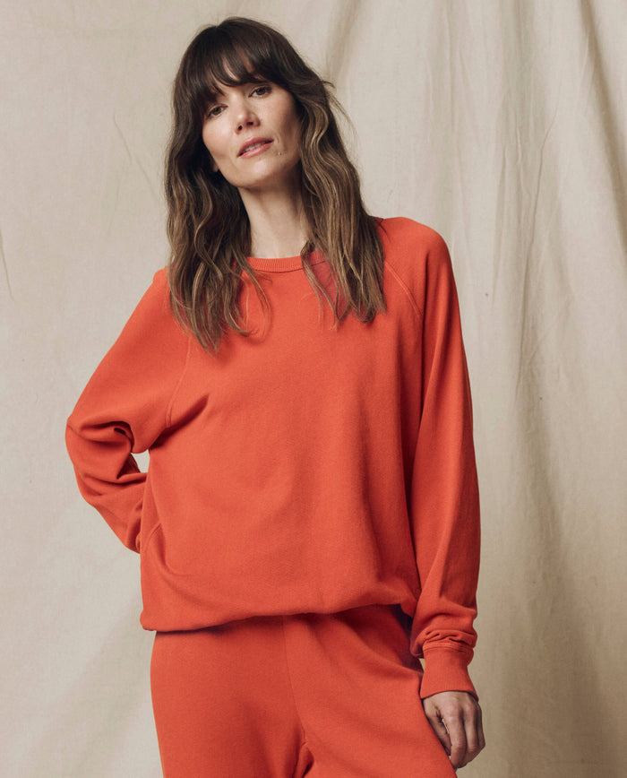 A woman with long brown hair wears THE GREAT.'s The College Sweatshirt in Solid Desert Clay from the SU25 SEASONAL KNITS collection, paired with matching pants, standing before a beige fabric backdrop.