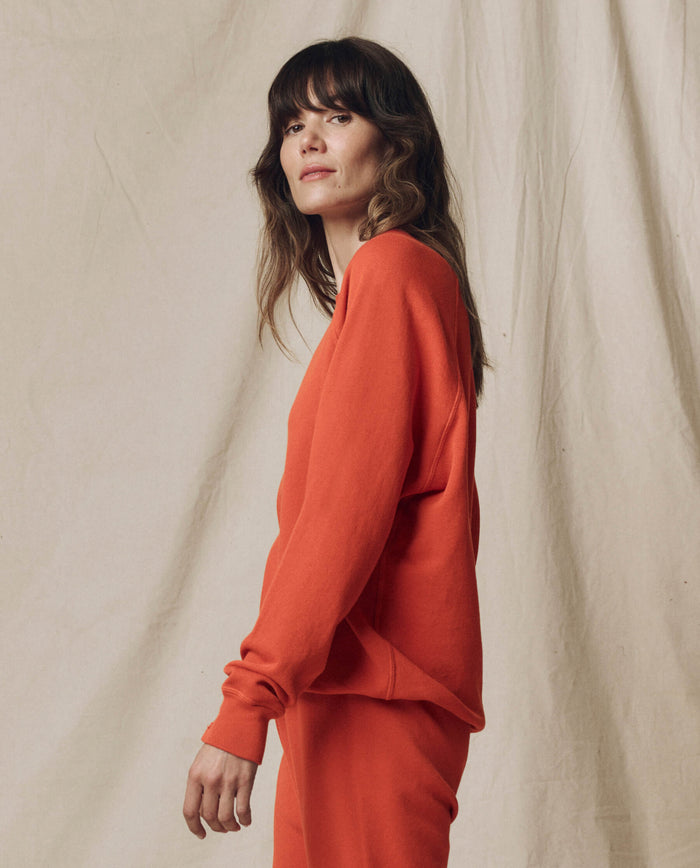 A woman with wavy brown hair stands sideways in front of a beige fabric backdrop, wearing THE GREAT.’s SU25 SEASONAL KNITS “The College Sweatshirt. Solid” in Desert Clay with matching pants.