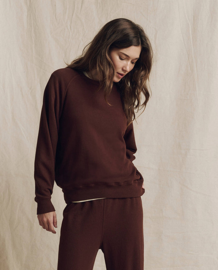Wearing THE GREAT.’s The College Sweatshirt in Solid Driftwood from the F26 SEASONAL CORE collection, a person stands against a beige fabric backdrop, looking downward with one hand by their side.