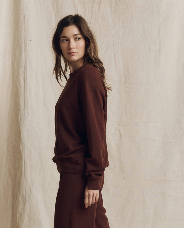 A person with long brown hair wears THE GREAT. The College Sweatshirt in Solid Driftwood from the F26 Seasonal Core collection, paired with matching pants, standing in front of a beige fabric backdrop and looking to the side.