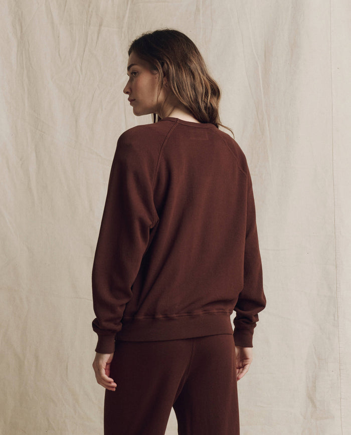 A person stands facing away against a beige fabric backdrop, showcasing THE GREAT.’s The College Sweatshirt in Solid Driftwood, styled with matching pants for a handcrafted Los Angeles-inspired look.